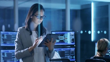 Aufkleber - Young Female Government Employee Wearing Glasses Uses Tablet in System Control Center. In the Background Her Coworkers are at Their Workspaces with Displays Showing Data. 