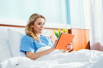 smiling woman using digital tablet while lying in hospital bed