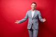 © luengo_ua - You are welcome! Cheerful young man in suit gesturing welcome sign and smiling while standing against red wall.