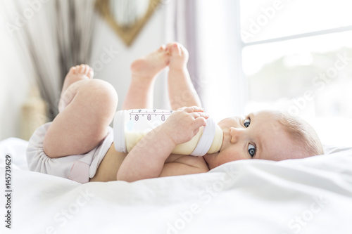 Fotografía Pretty baby girl drinks water from bottle lying on bed