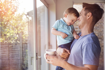 Father holds toddler son while drinking coffee at home, by the window