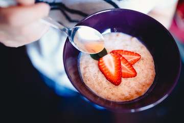 guy is holding a spoon with a strawberry cut with a knife in a plate with a porridge on the milk, two halves. The concept of cooking a tasty, healthy, healthy breakfast with oatmeal.