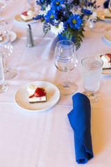 White tablecloth with wineglass, blue tissue napkin, Cheesecake with strawberry on the plate. Table set up