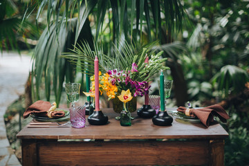 Wall Mural - A wooden table among tropical plants is served for two, on the table there are compositions of flowers and greens, candles and glasses