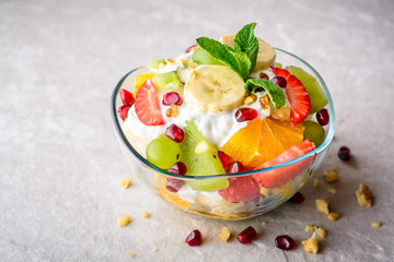 Fresh fruit salad with yogurt and walnuts in glass bowl on stone background.