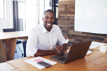 Young black man at desk with laptop computer looks to camera