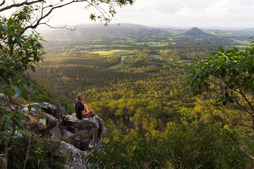 a young man sits, quietly meditating on top of a mountain with expansive views near noosa heads, aus