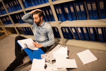 Tired businessman with scattered papers in file storage room 