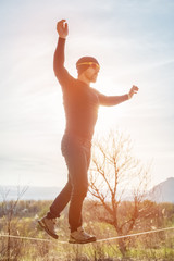 A man, aged with a beard and wearing sunglasses, balances on a slackline in the open air between two trees at sunset