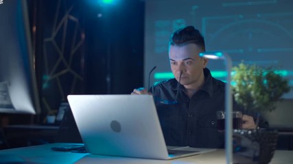 Tyred programmer or Developer sitting at laptop having coffee in modern office late at night