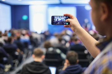 Audience at a business conference. Person taking photo with smart phone.