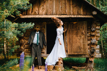 Young Beautiful Skinny Bride Girl In Vintage Wedding Dress And Hairstyle Standing Beyond Old Wooden House In Forest With Scarecrow From Fairytale. Classical Russian Fable Concept And Idea. Sorcery.