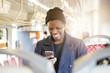 Laughing dark-skinned male in tram. African American using contemporary smartphone sitting in public transport, looking cheerful and happy reading funny stories browsing social network pages