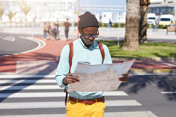 outdoor urban shot of serious dark-skinned european traveler in stylish clothing standing in the mid