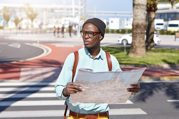 Handsome young Afro American tourist in eyewear and headdress having worried facial expression, looking for right direction using paper map while got lost in urban surroundings in foreign city