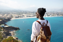 Back View Of Fashionable Young Male Backpacker Meditating On Top Of Mountain, Admiring Beautiful Nature Around Him. Unrecognizable Dark-skinned Male Looking At Blue Ocean From Birds Eye View
