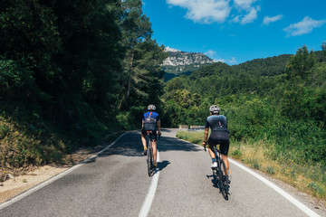 Wall Mural - Two male cyclists with shadows dressed in navy blue clothing racing, climbing a narrow mountian road high in Spain through green covered hillsides and sunny bright blue almost cloud free sky.