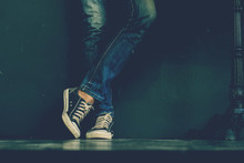 Young Fashion Man's Legs In Blue Jeans And Black Sneakers On Wooden Floor.