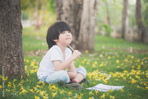 Little Asian Boy Sitting Under The Tree And Drawing In Notebook At