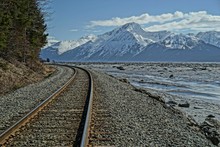 Train Track With View Of Water And Snow Covered Mountains.