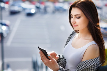 Young pretty stylish woman typing write message on smart phone in the street