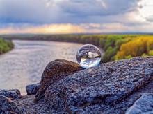 Magic Glass Ball On Old Stones. In The Background Is The Bend Of The River, Spring Forest And Cloudy Sky