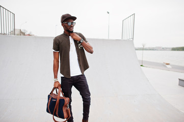 Portrait of sitting stylish african american man wear on sunglasses and cap with handbag outdoor against skate park. Street fashion black man.