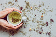 © fedorovacz - Tea background. Hands holding cup of hot tea. Dry herbal tea on the gray background, top view. Copy space