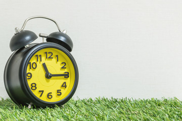 Wall Mural - Closeup black and yellow alarm clock for decorate show a quarter past eleven o'clock or 11:15 a.m. on green artificial grass floor and cream wallpaper textured background with copy space