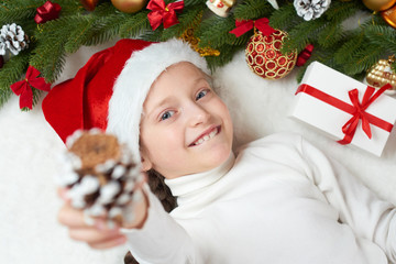 child girl having fun with christmas decoration, face expression and happy emotions, dressed in santa hat, lie on white fur background, winter holiday concept