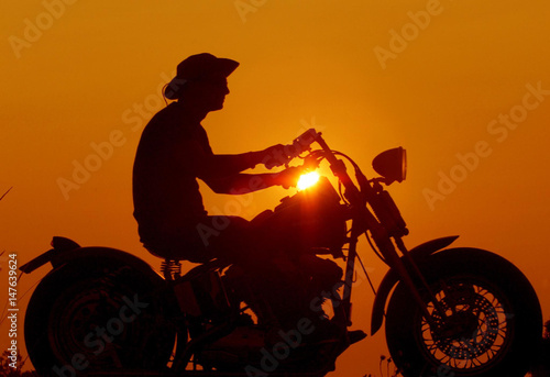 Cuadros en Lienzo A departing biker rides his old fashion Harley Davidson past a spectacular sunset on the last evenin