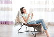 © Africa Studio - Young woman resting on modern chair and reading book at home in light room