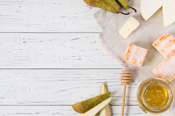 French soft cheeses from Normandy and Brittany regions sliced on cloth, honey, pear and spoon on white wooden background with copy space, top view