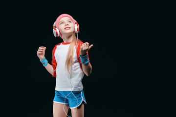 Wall Mural - Smiling little girl in sportswear wearing headphones and posing isolated on black, activities for children concept