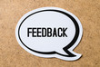 Feedback text in a speech bubble cut from paper or cardboard. High contrast picture with a light brown wooden cork board background. Useful icon styled design to website or social media for business. 