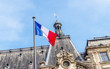Flag of France in front of an old building in Paris. Beautiful French architecture in historical city. Flag waving in the wind on a sunny day.