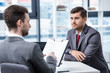 © LIGHTFIELD STUDIOS - Serious man in formal wear looking at businessman with clipboard during job interview, business concept