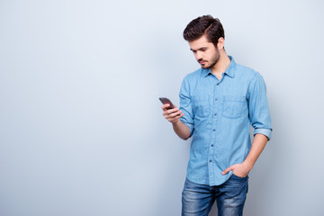 Sticker - Vertical portrait of young guy in jeans shirt, typing sms and holding his hand in pocket on pure light background