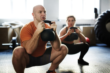 Young sporty woman and middle-aged bodybuilder doing kettlebell squat exercise during intensive training in gym