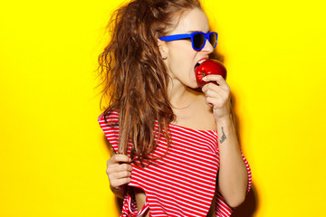 Beautiful young sexy girl in blue sunglasses and red striped T-shirt laughing and having fun and eating an apple on a yellow background