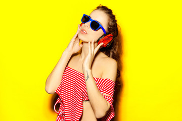 Wall Mural - Beautiful young sexy girl dj in blue sunglasses and red striped T-shirt laughing and having fun and listening to music in red headphones on a yellow background