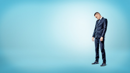 A sad businessman standing on blue background looking down with slumped shoulders.