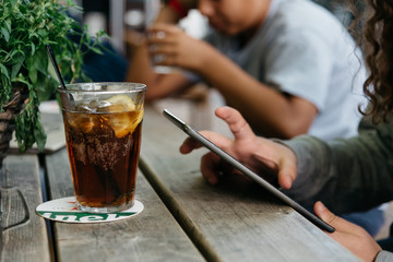 Refreshing glass of cola on wooden table in a bar while young is connected with tablet