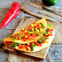 Wall Mural - Stuffed veggie omelette on wooden board, fork, knife on vintage wooden background. Homemade fried omelette stuffed with red and green bell peppers, canned corn and spices. Rustic stile