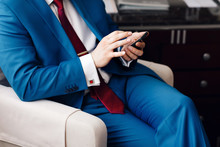 Businessman Keep A Phone In Hand Whilst Sitting On A Sofa In A Blue Suit. On Hand Expensive Mechanical Watch With Leather Strap. Shirt With Cufflinks