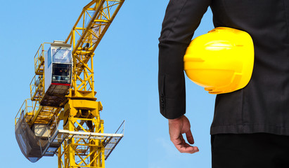 Wall Mural - businessman in suite with yellow safety helmet with construction crane tower against a blue sky in background,  business technology industrial concept.