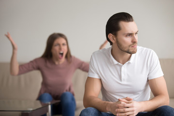 Young unhappy family couple quarrelling sitting apart at home. Upset frustrated serious husband thinking about problems sitting in foreground looking aside ignoring his hysteric nagging yelling wife