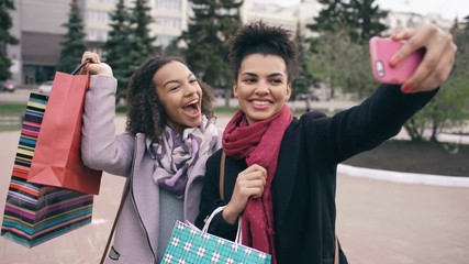 Wall Mural - Two cute african american woman taking selfie with shopping bags and smiling. Friends have fun after visiting mall sale.