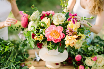 bouquet, people and floral arrangement concept - hands of two young woman drawing up a bouquet of beautiful roses, pink peonies, daisies in white vase, art workshop at wooden table in summer garden