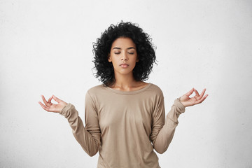 Consideration and praying. Beautiful calm young black female with Afro hairstyle keeping eyes closed while practicing yoga indoors, meditating, holding hands in mudra gesture, thinking about peace